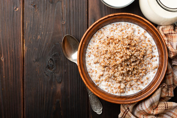 Buckwheat porridge with milk in ceramic bowl on dark wooden background. Top view, copy space.