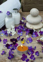 Obraz na płótnie Canvas Burning candle in the middle of purple pansy flowers and a spa set with skin lotion