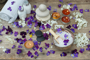 Obraz na płótnie Canvas Burning candles and spa treatment kit with bath salt, stones for hot massage and skin lotions in the middle of purple flowers of pansies