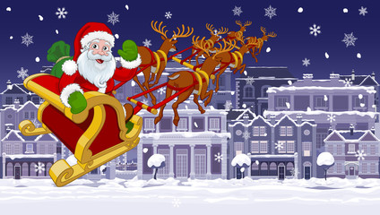 Christmas night street scene with Santa Claus in his Christmas sleigh with reindeer flying over Victorian style houses, shops and other buildings in the snow.