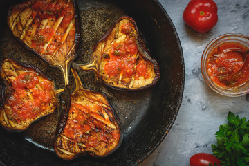 Grilled glazed eggplants on black pan or baking sheet with red sauce, parsley, tomatoes on grey background. Copy text area for menu design. Horizontal top view. Healthy vegetarian snacks concept.
