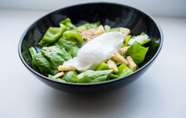Salad of rucola leaves, green bell pepper, flax seeds, hard cheese, poached egg in a black plate on a light background.