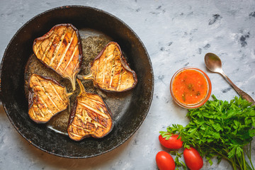 Grilled glazed eggplants on black pan or baking sheet with red sauce, parsley, tomatoes on grey background. Copy text area for menu design. Horizontal top view. Healthy vegetarian snacks concept.
