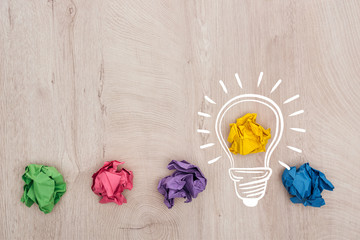 top view of multicolored crumpled paper balls and light bulb illustration on wooden surface, business concept