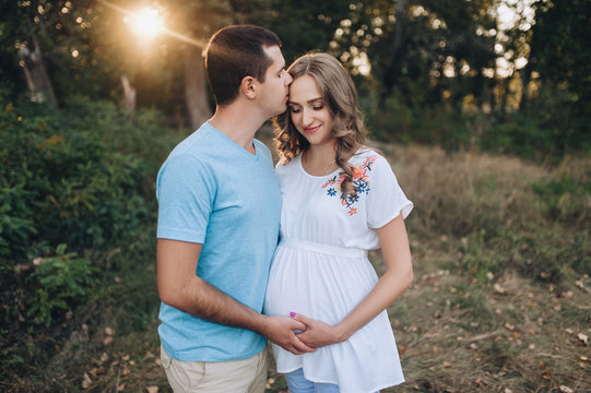 A loving man hugs and kisses a beautiful pregnant blonde woman at sunset in the forest. Portrait of a young family expecting a baby with a ray of sunshine. Pregnancy picture, concept.