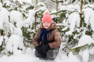 Fototapeta na wymiar Happy little girl in winter clothes sitting outdoor under spruce tree covered with snow