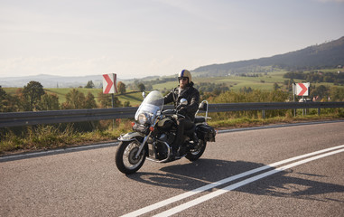 Bearded biker in helmet, sunglasses and black leather clothing riding cruiser motorcycle along sharp turn of empty road on bright summer day on misty background of rural landscape and distant hills.