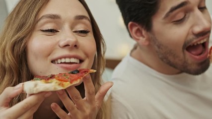 Attractive happy couple man and woman lying on bed at home and enjoy eating tasty pizza 