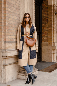 street style portrait of an attractive woman wearing a beige trench coat, denim jeans, black ankle boots and brown leather bag, crossing the street. fashion outfit perfect for autumn fall winter