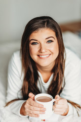 Interior portrait of happy young woman relaxing in bedroom, wearing pajamas, holding cup of hot chocolate