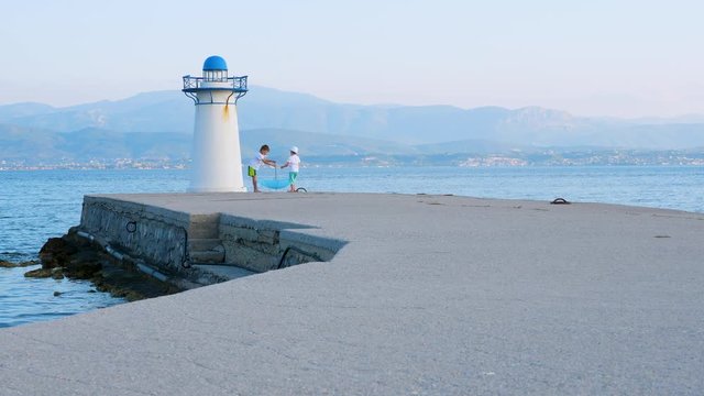 Two cheerful boys playing with blue umbrella on pier with lighthouse. White lighthouse on dock in island Evia, Greece. Positive emotions while traveling to Greece. Traveling with kids