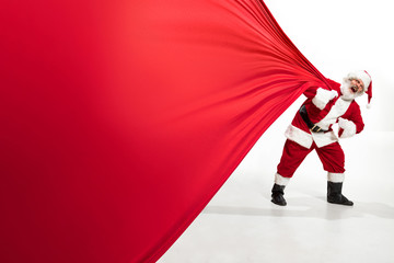 Santa Claus pulling huge bag full of christmas presents isolated on white background. Caucasian male model in traditional costume. New Year 2020, gifts, holidays, winter mood. Copyspace for your ad.