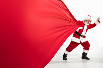 Fototapeta na wymiar Santa Claus pulling huge bag full of christmas presents isolated on white background. Caucasian male model in traditional costume. New Year 2020, gifts, holidays, winter mood. Copyspace for your ad.
