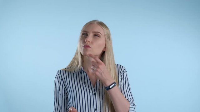 Blonde young woman holding her chin with hand while thinking about something on blue background. She looking at smartphone and making plans.