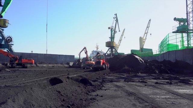 Coal Marine Terminal. Shooting a general plan. A red bulldozer picks up black coal to a crane that loads the mineral onto a marine cargo ship.