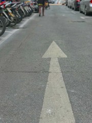 The guy is walking on the road with white traffic arrow pointing forward.