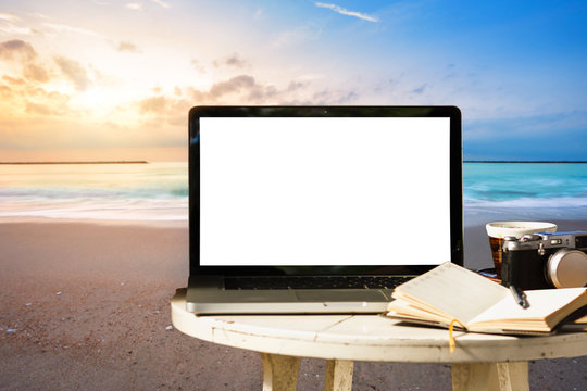 Mockup of laptop computer with empty screen with camera,notebook,coffee cup on table at landscape early sunrise over blue the sea background,working on the beach,Freelance work and holiday traveler.