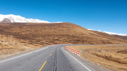 Centered view of bend asphalt road in plateau of northwest China, with autumn pasture and snow mountains background.
