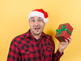 Happy surprised man in a red hat holding a gift with admiration on his face. Positive human emotions at christmas. Yellow background, copy space.