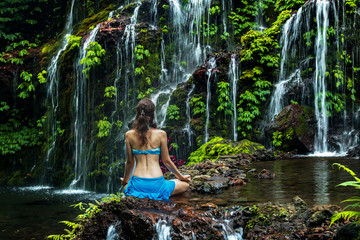 Young woman sitting on the rock, practicing yoga near waterfall. Hands in gyan mudra. Banyu Wana...