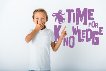 shocked kid covering face while looking at camera and pointing with finger at time for knowledge letters on white
