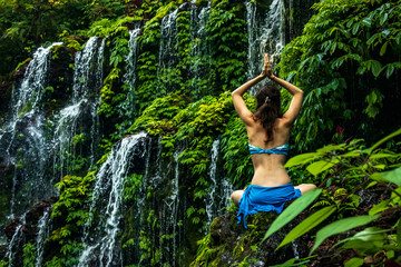 Woman sitting on the rock, practicing yoga. Young woman raising arms with namaste mudra near waterfall. Banyu Wana Amertha waterfall, Bali. View from back.