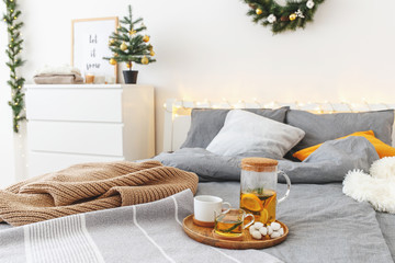 Christmas cozy winter home decor. New year interior decorations. Bed with grey linen, blanket, pillows, plaid, christmas tree, led garland light, citrus tea on wooden tray. White stylish bedroom.