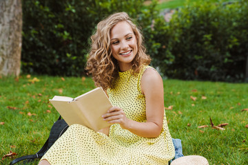 Image of pleased blonde woman smiling and reading book in green park