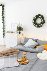 Christmas cozy winter home decor. New year interior decorations. Bed with grey linen, blanket, pillows, plaid, christmas tree, wreath, led garland light, citrus tea on tray. White stylish bedroom.