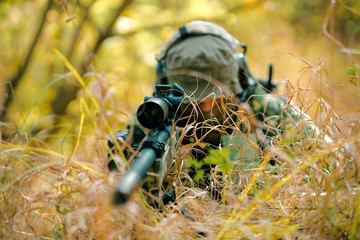Closeup man in uniform with sniper rifle, front view. Airsoft soldier lay in long grass and check...