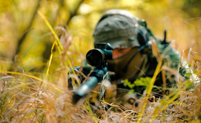 Closeup man in uniform with sniper rifle, front view. Airsoft soldier lay and aims at the sight in long grass