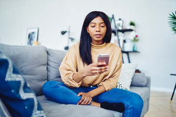 Dark skinned hipster girl installing new app on smartphone while spending free time at home sitting on sofa,thoughtful African American woman making booking online on browsed website via mobile phone