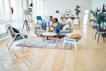Happy african american young family sitting on sofa in modern designer living room in apartment,...