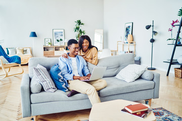 Cheerful african american wifie looking at her husband working on freelance via laptop computer at home interior, dark skinned romantic couple booking tickets for trip vacation together via netbook.