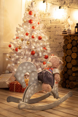 Beautiful Christmas composition: a vintage children's wooden-swing wooden horse with a Teddy bear, a mediocre Christmas tree with red balls and gift boxes, an old brick wall and a burning garland.