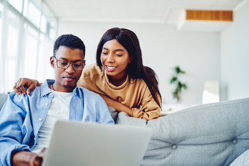 Smiling african american woman looking at freelance job of husband sitting with laptop computer at sofa in living room, positive dark skinned couple in love making shopping together  in web store.