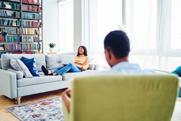 Fototapeta premium Happy african american woman laughing lying on sofa during free time at modern home interior room, back view of dark skinned man sitting in chair looking at his wife enjoying relaxing on couch.
