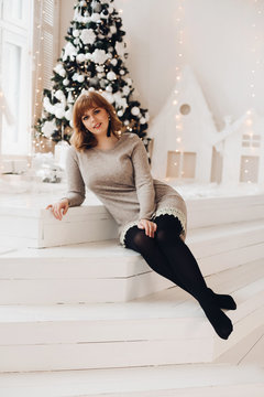 Full length portrait of attractive chestnut-haired woman pondering away in her grey dress and earrings. She is sitting on white wooden steps in front of decorated Christmas tree. Snowflakes fall down.