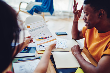 Cropped view of African American colleagues brainstorming on accounting report sitting at desktop with stationery, dark skinned man studying with tutor holding graphs with infographic statistics