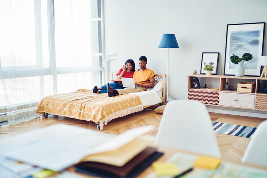 Young african american couple in love recreating on cozy bed making shopping on home goods web page using laptop computer, smiling dark skinned marriage watching movies on netbook on free time  .