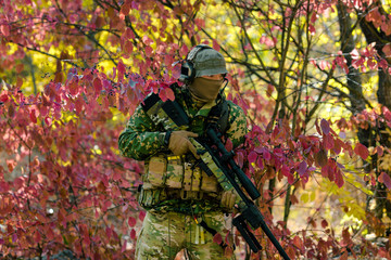 Naklejka premium Man in green uniform with sniper rifle. Airsoft soldier stand in red bush. Horizontal photo