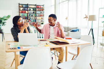 Cheerful african american colleagues excited with new successful ideas for startup sitting together at table, happy dark skinned guy explaining his strategy to pensive woman discussing opinions.