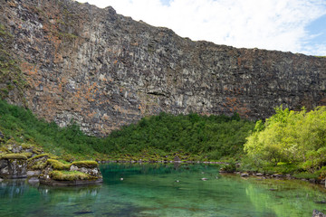 Clear glacial lake under a tall rocky hill wall, Iceland Asbyrgi