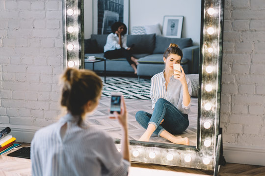 Charming young woman in casual wear sitting on floor in front of stylish mirror and making photo on modern smartphone device.Gorgeous hipster girl taking picture for blog on cellular resting at home