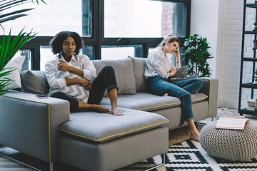 Obraz premium Offended african american young woman ignoring live communication with best caucasian friend after conflict sitting at home interior.Stubborn hipster girl with dark skin don't talk with friend