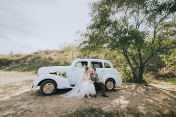 Fototapeta na wymiar Beautiful newlyweds are sitting and hugging near an old retro car and summer nature. Wedding portrait of a stylish, smiling groom and lovely bride with curly hair. Photography and concept.