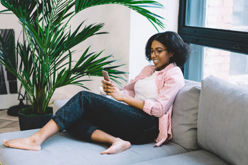 Positive female blogger with dark akin laughing during chatting with friends in network on modern smartphone.Cheerful african american young woman doing shopping on website on cellular resting at home