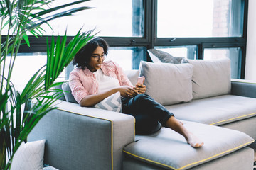 Obraz premium Smiling african american young woman laughing while watching funny video in blog on smartphone device relaxing on comfortable couch in modern apartment.Positive hipster blogger chatting on cellular