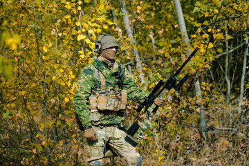 Airsoft man in uniform hold sniper rifle on yellow forest backdrop. Side view