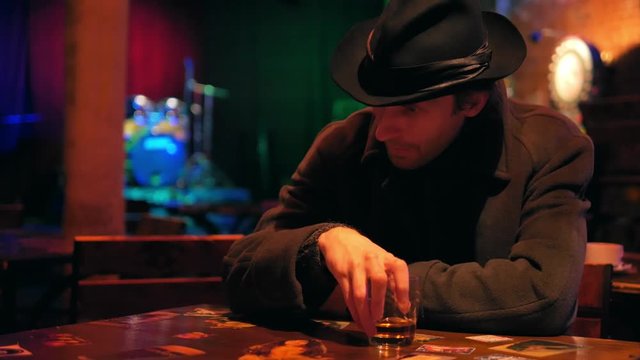 Brutal man in a hat with a glass of whiskey sits alone in an empty bar
